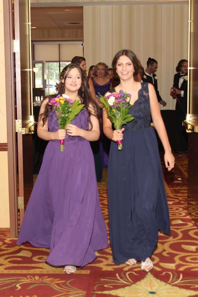 Ella in a blue gown and Alexus in a purple gown lead the wedding party with Andrew and Jarrod in the background during the grand march, captured by Betsy Murphy.