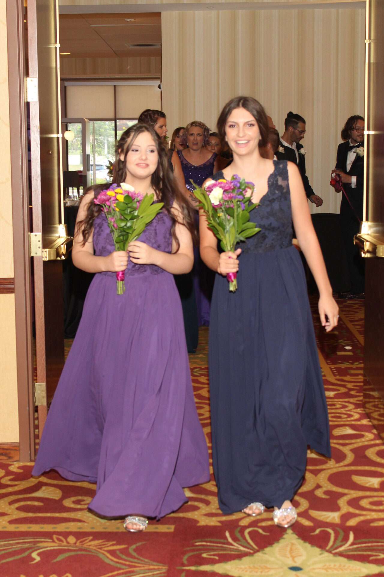 Ella in a blue gown and Alexus in a purple gown lead the wedding party with Andrew and Jarrod in the background during the grand march, captured by Betsy Murphy.