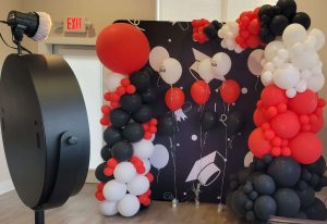This picture shows balloons decorations and a beauty mirror photo booth by GBF Bespoke Balloons and Event Services. The balloons are red, black, and white. There is an organic half balloons archway and a beautiful balloon column that matches with a giant 3' red balloon atop the column.