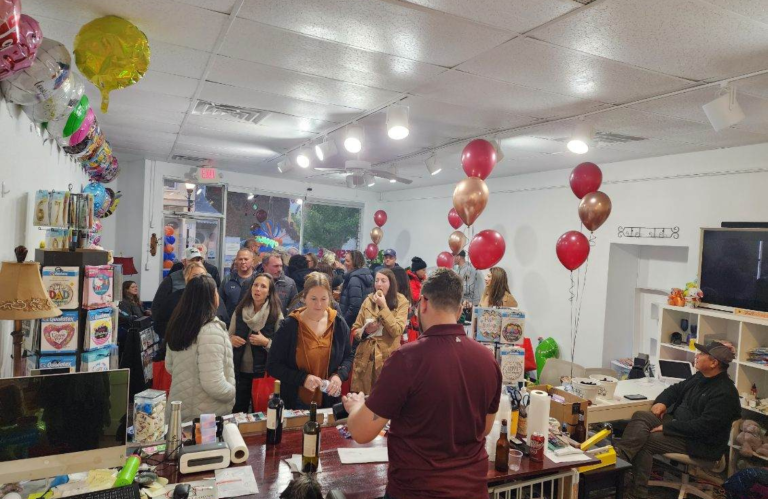 Interior view of GBF Bespoke Balloons and Event Planning Center during Wauktober. The store is alive with guests milling around. A prominent figure, Jarrod, in a maroon shirt stands behind a counter, pouring wine and beer for attendees. Above, an array of colorful balloons decorates the ceiling, while various balloon-themed merchandise and party essentials line the shelves to the left. On the right, a TV screen displays a man's face, and nearby, another individual sits comfortably, watching the scene unfold. The atmosphere appears festive and welcoming.