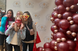 Guests at Waukesha Civic Theatre posing with props in front of GBF's Beauty Mirror, accompanied by a burgundy demi arch balloon display.