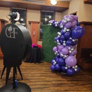 GBF's beauty mirror photo booth stands beside a demi arch of lilac, violet, and chrome silver balloons, ready for Pam's retirement party photos.