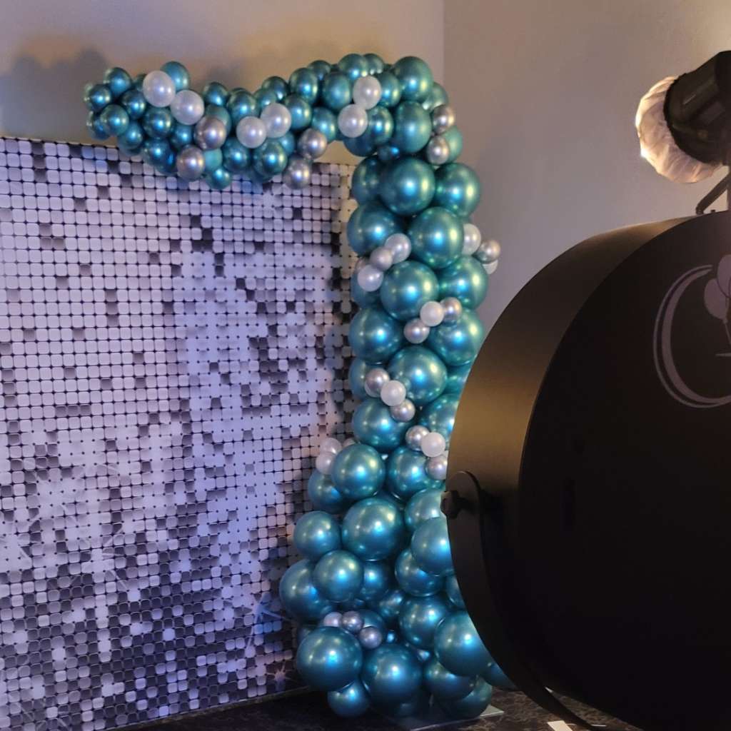 An elegant Beauty Mirror Picture Booth stands beside a stunning organic demi archway made of lustrous teal and pearl balloons, designed to add a touch of sophistication to any celebration.