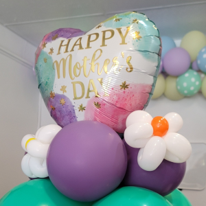 An air-filled balloon decor piece for Mother's Day, featuring a heart-shaped 'Happy Mother's Day' mylar balloon amid a cluster of elegant purple and teal latex balloons with a handmade balloon flower.