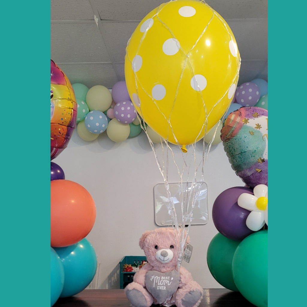 A plush 'Best Mom Ever' teddy bear paired with a vibrant yellow helium balloon encased in a decorative net, creating a charming balloon bouquet.