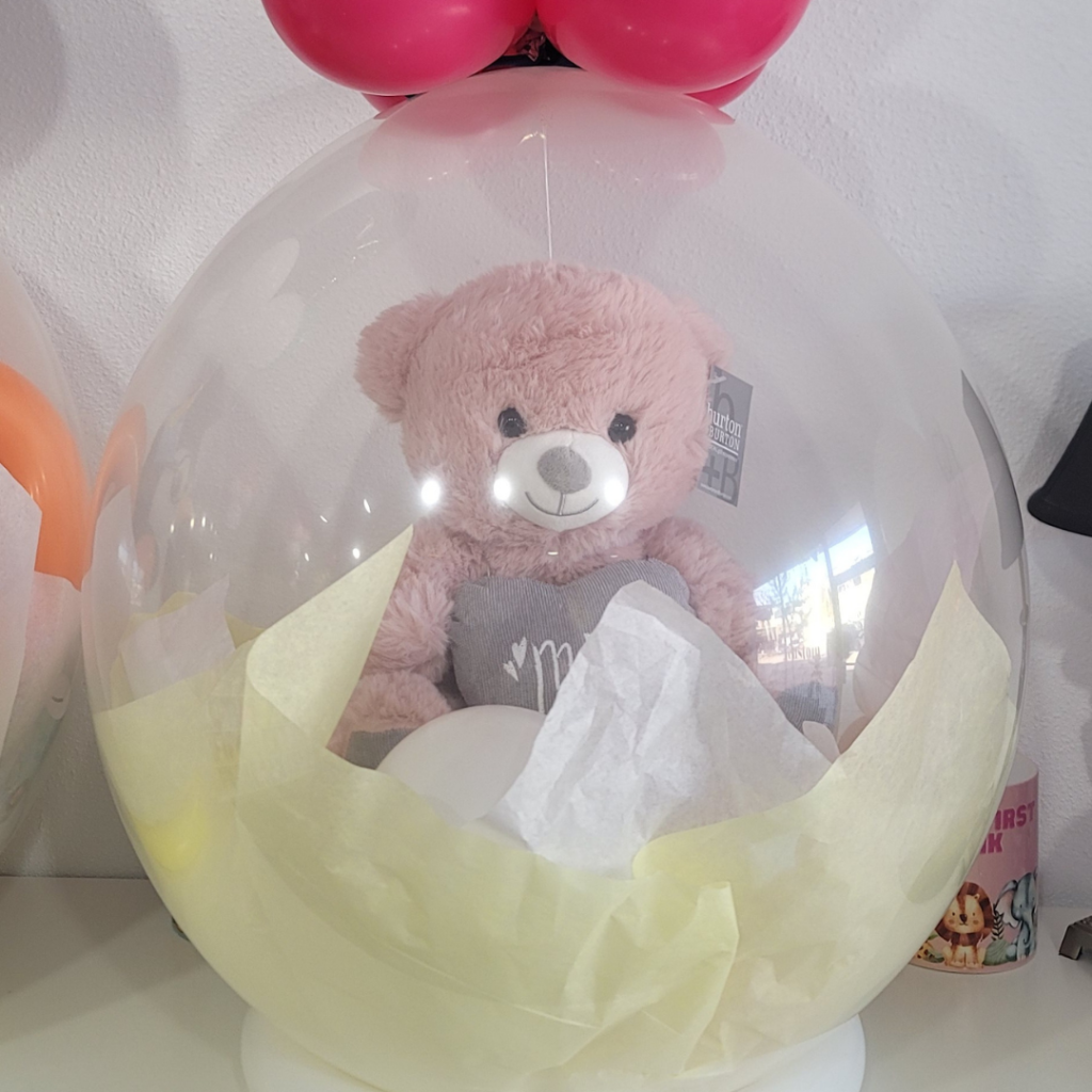 Close-up view of a cute pink teddy bear with a 'Best Mom Ever' shirt, encased within a clear stuffed balloon, adorned with a yellow tissue paper.