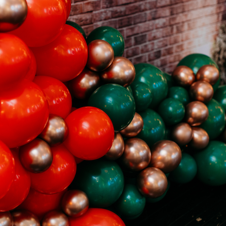A lively organic balloon wall arrangement in festive red, green, and metallic colors.